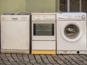 Old white dishwasher, electric stove, and washing machine lined up on a cobblestone street for disposal.
