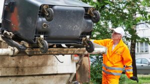 Sanitation worker in high-visibility orange uniform operating a rear-loading garbage truck to empty a large wheeled trash bin.