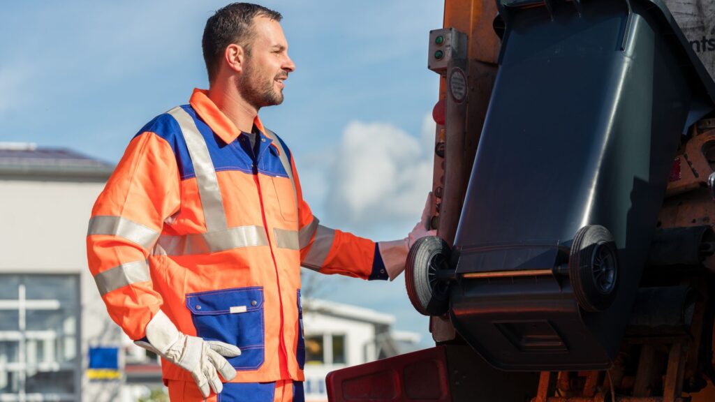 Smiling sanitation worker in an orange and blue high-visibility safety jacket operating a mechanical lift to empty a black trash bin.
