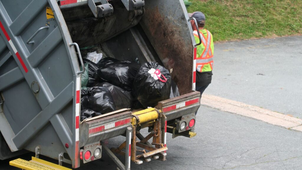 Sanitation worker in a high-visibility safety vest standing next to a rear-loading garbage truck filled with black trash bags on a residential street.