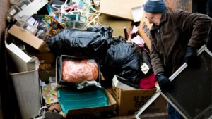 Worker loading a metal window frame into a large dumpster filled with household junk, cardboard egg boxes, and black trash bags.