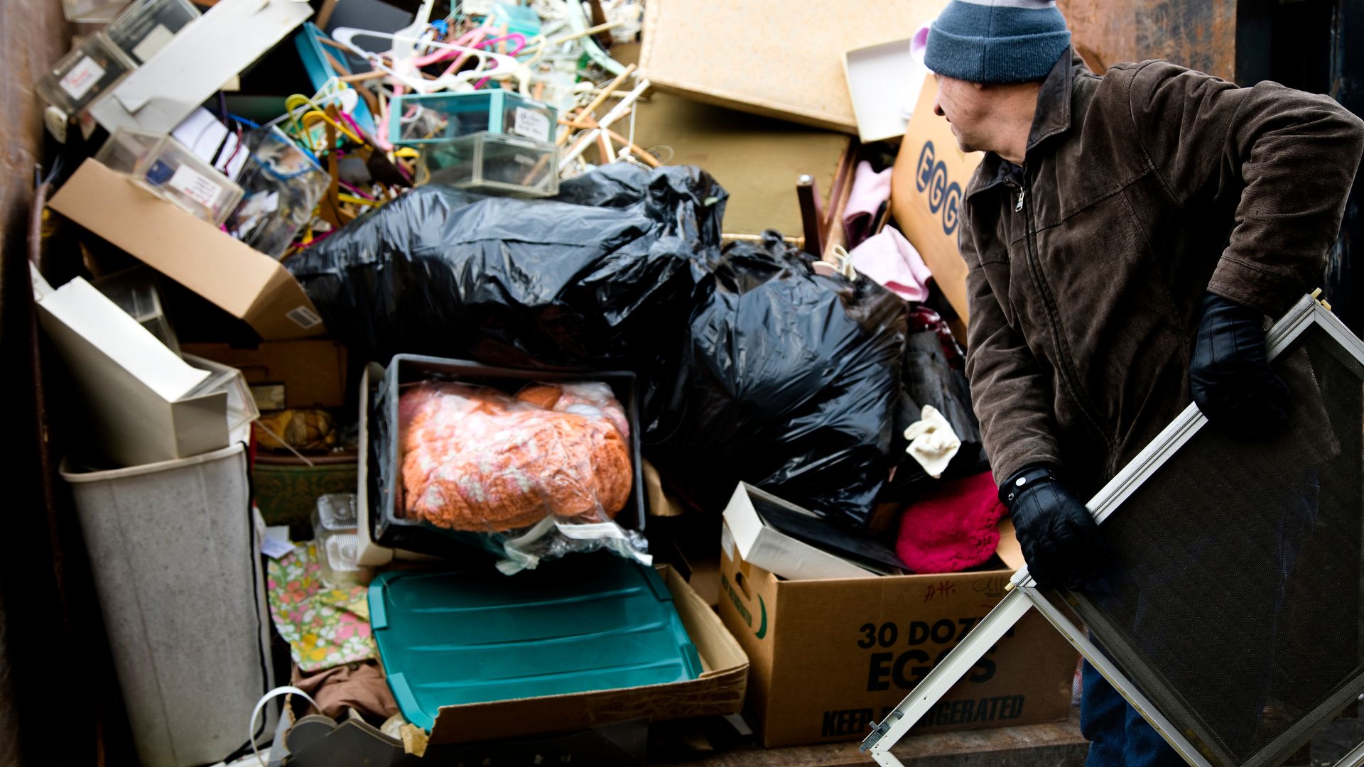 Worker loading a metal window frame into a large dumpster filled with household junk, cardboard egg boxes, and black trash bags.