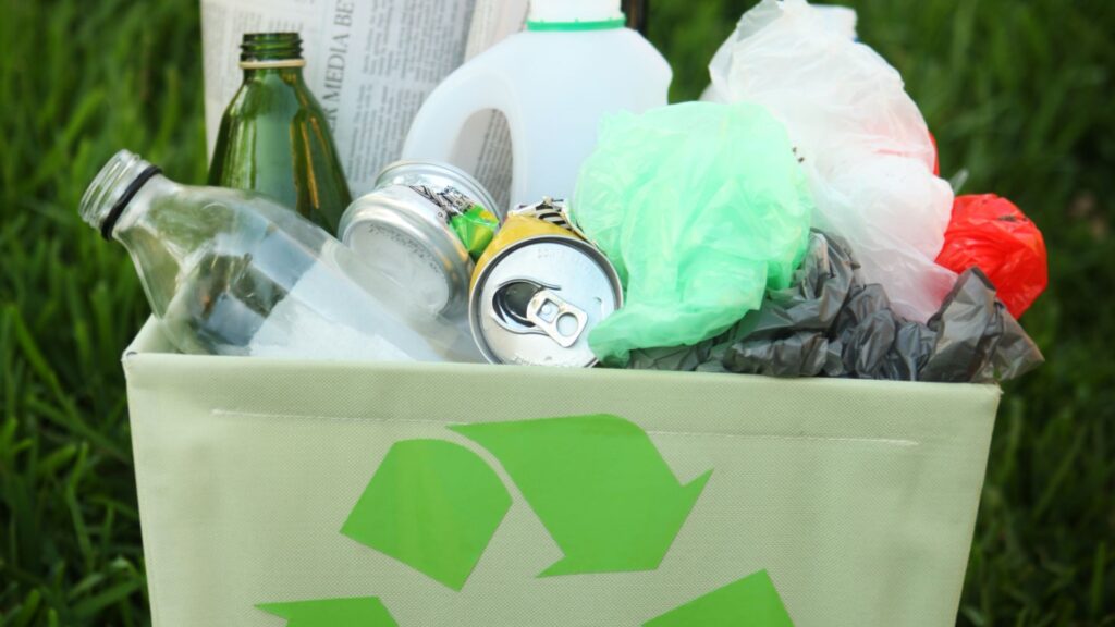 Green recycling bin overflowing with sorted glass bottles, aluminum cans, plastic jugs, and bags on a grass background.